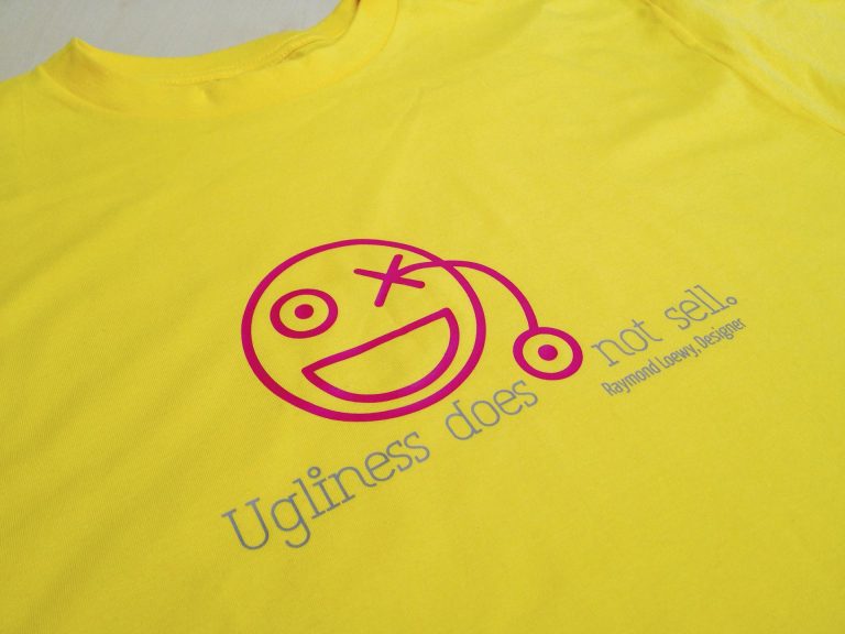 Gelbes T-Shirt mit pinkem Smiley und dem Zitat „Ugliness does not sell. – Raymond Loewy, Designer“ in blauer Schrift.