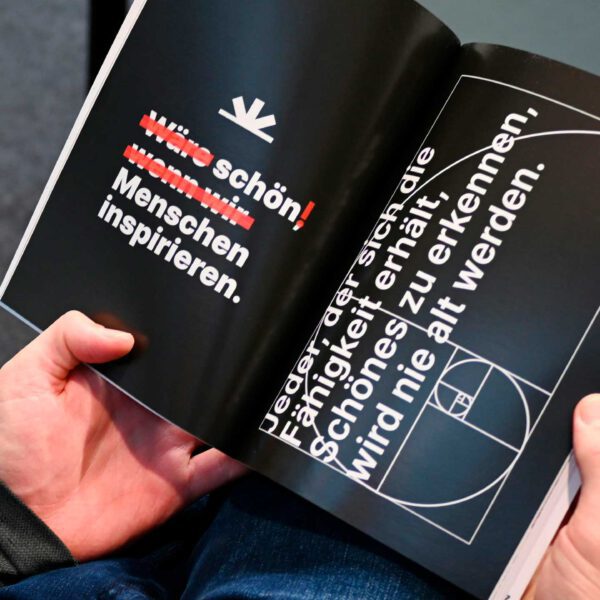 Person hält eine aufgeschlagene Design-Broschüre mit modernem, typografischem Layout und Zitat von Franz Kafka.
