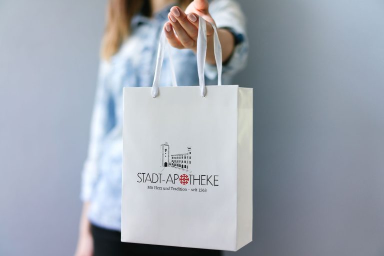 Eine Person hält eine weiße Papiertüte mit dem Logo der "STADT-APOTHEKE Mühldorf" und dem Slogan "Mit Herz und Tradition – seit 1563" vor einem neutralen grauen Hintergrund.