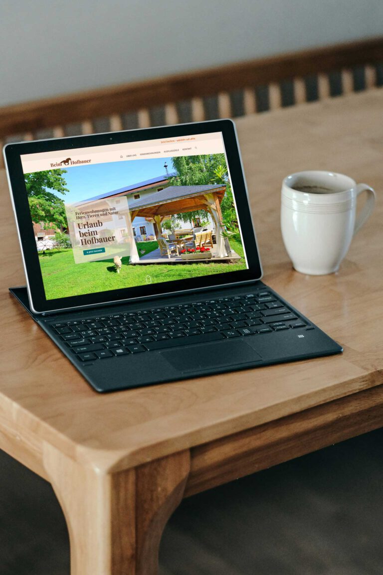 Tablet auf einem Holztisch mit geöffneter Webseite der Ferienwohnungen Beim Hofbauer, daneben steht eine weiße Kaffeetasse.Auf einem Holztisch steht ein Tablet mit Tastatur, auf dessen Bildschirm die Startseite der Ferienwohnungen Beim Hofbauer zu sehen ist. Die Webseite zeigt ein Gartenmotiv mit Pavillon und Sitzgruppe. Neben dem Tablet befindet sich eine weiße Kaffeetasse.
